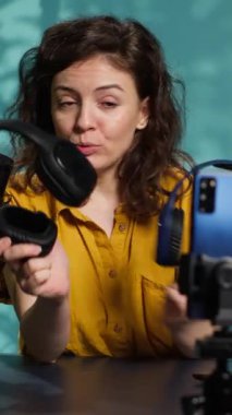 Seyircilere oyun kulaklığı sunan loş ışıklı ev stüdyosunda internet kanalı için dikey video ses mühendisi video kaydı. İzleyicilere müzik dinleme cihazı gösteren kadın, kamera A