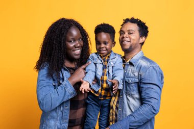 Stüdyoda sarı arka plana gülümseyen Afro-Amerikan ailesi sevgi ve içten şükranla poz veriyor. İyimser anne ve baba, tatlı oğullarını kucaklıyor, birlikte vakit geçiriyorlar..