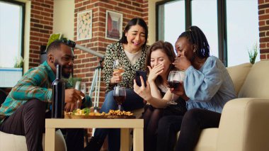 Mezunlar toplantısındaki çok ırklı arkadaşlar akıllı telefon ekranında çocukluklarından komik videolar izliyorlar. Oturma odasında neşeli konuklar cep telefonundan gençliklerinden klipler izliyorlar.