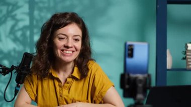 Stüdyoda gülümseyen bir kadın telefonuyla video kaydediyor, aboneleri selamlıyor. Mutlu çevrimiçi yıldız Q 'yu sunar ve izleyicilerle cep telefonu, selamlama hayranları ve kamera A' yı kullanır.