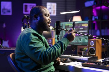 Genç sanatçı, akıllı telefonundan mısralar okuyarak ve mikrofonla şarkı söyleyerek ev stüdyosunda onun tarafından yazılan yeni bir şarkıyı kaydediyor. Ses mühendisi ses efektlerini düzenlemeye hazırlanıyor.