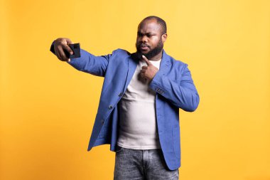 Kendine güvenen Afro-Amerikan bir adam akıllı telefon kullanarak selfie çekiyor ve çenesini işaret ediyor. Kibirli insan cep telefonu fotoğraf makinesi, narsisizm konsepti, stüdyo arka planı kullanarak fotoğraf çekiyor.