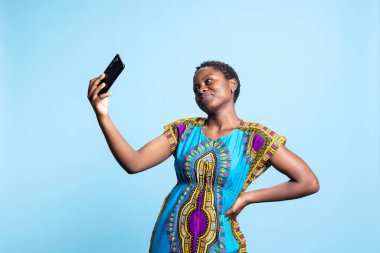 Akıllı telefon uygulamasında fotoğraf çeken enerjik mutlu kadın, iyimserliğini ve sosyal medya platformlarında büyüleyici gülümsemesini gösteriyor. Afrikalı Amerikalı kız cep telefonuyla fotoğraf çekiyor, doğal güzellik..