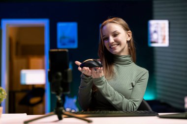 Neşeli genç cep telefonunu tripodla internet kanalı için video çekmek için kullanıyor, kablosuz fareyi sunuyor. Mutlu kız cep telefonuyla bilgisayar çevresel aygıtı incelemesi kaydediyor