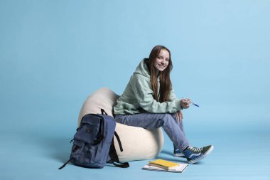 Sırt çantalı ve okul defterli gülümseyen kadın portresi stüdyo arka planında izole bir şekilde duruyor. Okul malzemeleriyle neşeli kız rahatlıyor, üniversiteye gitmeye hazırlanıyor.