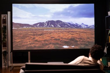 Adam kanepeye uzanmış, modern video projektöründe güzel bir sinematografi ile seyahat belgeseli izliyordu. Ev sinemasında dağlık manzaranın tadını çıkaran kişi.