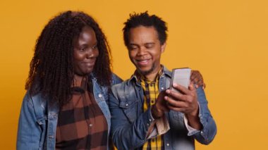 Afro-Amerikan genç çifti akıllı telefondaki fotoğraflarla birlikte eğleniyor ve birlikte hoş vakit geçiriyorlar. Tatlı karı koca cep telefonuyla fotoğraf çekiyor. Kamera A.
