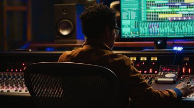 Afrika kökenli Amerikalı ses tasarımcısı, hareket eden faders ve renkli sayaçlarla ses konsolu kullanarak profesyonel stüdyoda şarkıları karıştırıyor ve ustalaştırıyor. Müzik yapımcısı kontrol odasında çalışıyor. Kamera A.