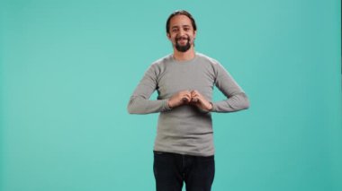 Sevinçli, dost canlısı bir adamın portresi. Elleriyle kalp sembolü hareketi yapıyor. Gülümseyen ve sevgi gösteren kişi, mavi stüdyo arka planında izole edilmiş, kamera A