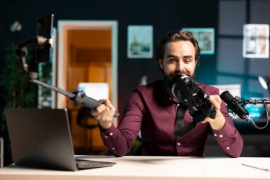 Teknoloji vlogcusu stüdyo dairesindeki VR gözlüklerini inceliyor, özellikleri sunuyor, telefonla kayıt yapıyor. Teknoloji hayranları için selfie çubuğunda cep telefonu kullanan bir adam sanal gerçeklik kulaklığı incelemesi yapıyor.