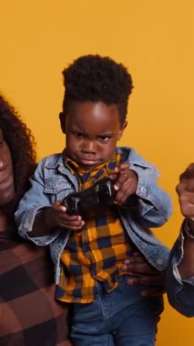 Dikey Video Neşeli Afro-Amerikan babası oğluyla konsolda video oyunu oynuyor, kumandayı sarı arka planda eğlenmek için kullanıyor. Mutlu, genç bir aile keyfine bakıyor.