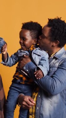 Stüdyoda genç bir çocukla birlikte Afrikalı Amerikalı bir ailenin dikey video portresi birbirlerine sevgi ve minnettarlık sunuyor. Aptal, sevgi dolu ebeveynler, küçük çocuklarını güldürmek için oynaşıyorlar.