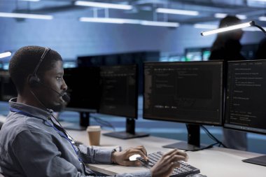 Çağrı merkezinde çalışan çalışanlar, veri merkezi işletmeleri için dış kaynak hizmetleri sağlıyorlar. İş yerindeki Afrikalı Amerikalı adam sunucu merkezindeki saha teknisyenleriyle mikrofonla konuşuyor.