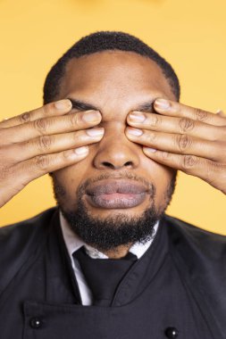 Restaurant worker showing the three wise monkeys gesture in studio, covering his eyes and see no evil as a sign of wisdom and concealment. Butler in apron shows wise symbolic metaphor. clipart