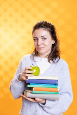 Portrait of radiant woman holding pile of books and organic apple, conveying joy of reading and eating healthy, studio background. Upbeat lady with novels and fresh fruit, harmonious lifestyle concept clipart