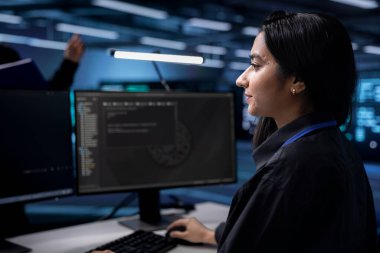 Indian technician in data center updating systems to keeps performance up and protect servers from exploitable software vulnerabilities. Woman looking for errors causing equipment to slow down clipart