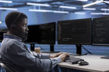 Veri merkezindeki mühendis çağrı merkezi görevlerini yönetiyor, birden fazla bilgisayar ekranını izlerken kulaklığa konuşuyor. Afrikalı Amerikalı adam kulaklıkla müşterilerle konuşuyor.