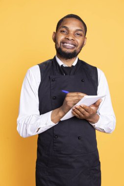 Profesyonel yemek şirketi müşterilerine fatura için not defteri hazırlıyor. Sarı arka planda istenen yiyecekleri not alıyor. Önlüklü garson, müşteri memnuniyeti için defter servisi..