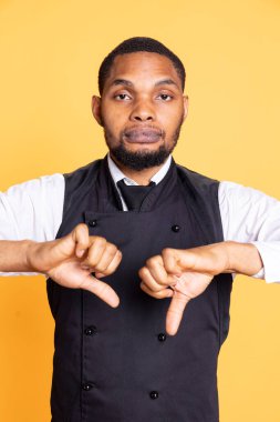 Yemek endüstrisi çalışanı önlüklü, sarı arka plana karşı olan anlaşmazlığını gösteren baş parmak işareti gösteriyor. Üniformalı, hoşnutsuz uşak, sembol göstermiyor..