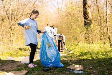 Neşeli, gururlu genç bir çocuk ormandaki çöpleri temizlemeyi bitiriyor, doğal çevreyi restore ediyor ve orman kirliliğine yardım ediyor. Plastik atıkları geri dönüştürüyor ve uzun pençe aracı kullanıyor.