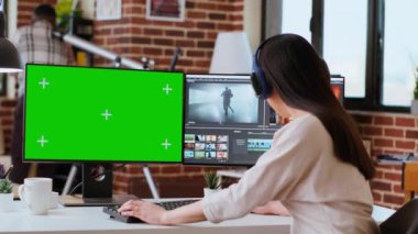 Modern bir mekanda yeşil ekranın yanında çalışan profesyonel Asyalı kadın çift monitör ve gelişmiş video kurgu yazılımı kullanıyor. Yapım sonrası çoklu görev. Kamera A.