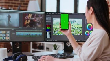 Modern bir mekanda yeşil ekranın yanında çalışan profesyonel Asyalı kadın çift monitör ve gelişmiş video kurgu yazılımı kullanıyor. Yapım sonrası çoklu görev. Kamera A.