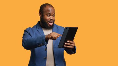 Sosyal medya yayınını kontrol etmek için tablet dokunmatik ekranda mutlulukla kayan bir adam. Afro-Amerikalı kişi internet sitelerine göz atıyor, içerik, stüdyo arka planı, kamera A ile eğlendiriliyor.