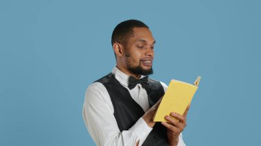 Genç kapıcı, stüdyo molasında kitap okuyor. Smokinli otel görevlisi, mavi arka planda. Belboy tatil köyü personeli edebiyat ve konferansı sever. Kamera A.