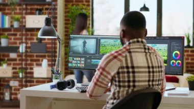 Erkek video editörü, oturma odasındaki modern ev ofisinden çifte monitör kurulumu ve düzenleme yazılımı ile birden fazla iş yapıyor. Medya üretimi için üretim sonrası görevler tamamlanıyor. Kamera B.