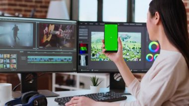 Modern bir mekanda yeşil ekranın yanında çalışan profesyonel Asyalı kadın çift monitör ve gelişmiş video kurgu yazılımı kullanıyor. Yapım sonrası çoklu görev. Kamera A.