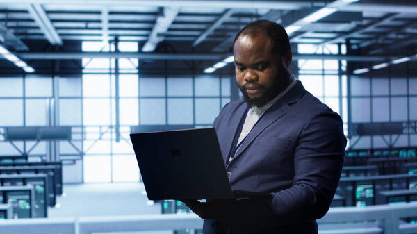 African american IT operator doing maintenance using laptop to prolong data center equipment life span. IT professional in server hub workspace monitoring server mainframes using notebook, camera A