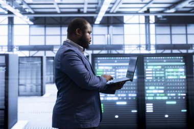 Employee in server hub using notebook device to monitor energy consumption across parts. IT professional in data center making sure infrastructure rigs are functioning optimally clipart