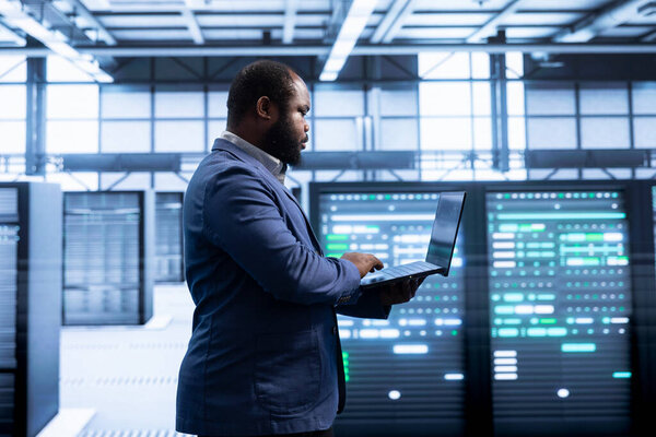 Employee in server hub using notebook device to monitor energy consumption across parts. IT professional in data center making sure infrastructure rigs are functioning optimally