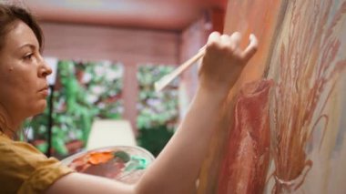 Dişi ressam stüdyoda bir sanat eseri yaratmak için bir tuval üzerinde çalışıyor, kılları kullanarak hassas bir şekilde boya uyguluyor. Sanat galerisi için eşsiz bir resim yapma becerisini sunuyor. Kamera B.