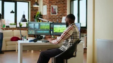 Erkek video editörü, oturma odasındaki modern ev ofisinden çifte monitör kurulumu ve düzenleme yazılımı ile birden fazla iş yapıyor. Medya üretimi için üretim sonrası görevler tamamlanıyor. Kamera A.