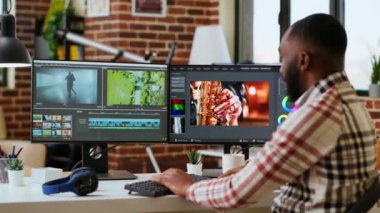 Afrikalı Amerikalı serbest çalışan, evinde arka plan videosunu düzenliyor, çift ekran klipleri hazırlıyor. Profesyonel adam, yaratıcı ve multimedya projeleri için teknolojik araçlar kullanır. Kamera B.