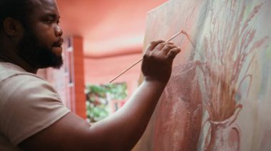 Erkek sanatçı suluboya ve palet ile modern bir sanat eseri çiziyor, sanat ve zanaat atölyesinde yeni eseri üzerinde çalışıyor. Genç ressam tuvalde kırsal bir başyapıt yaratıyor. Kamera B.