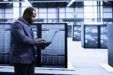 Bilgisayar bilimci veri merkezinde yürüyor, yönlendiriciler, anahtarlar ve güvenlik duvarları gibi ağ altyapısını yapılandırmak için dizüstü bilgisayar kullanıyor. Sunucu çiftliğinde teknik sorunlar arayan programcı