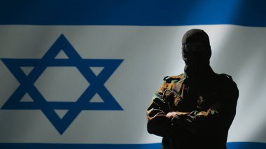 Maske takan Mossad operatörü İsrail bayrağı önünde gururla duruyor ve ülkesine hizmet etmekten gurur duyuyor. İsrail gizli ajanı kumanda odasında çapraz silah, kamera B
