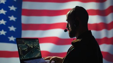 A.B.D. Hava Kuvvetleri harekat subayı cihazdaki izleme iletimini inceliyor, kontrol odasından yer askerlerine kulaklık kullanarak veri aktarıyor, ABD bayraklarından izole edilmiş, kamera B