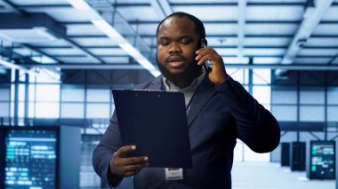 Bilgi işlem uzmanı veri merkezinin kontrol listesi evraklarını inceliyor, sunucu çiftliğinde telefonla konuşuyor. Afrika kökenli Amerikalı adam, sunucu platformlarıyla çevrili, telefon görüşmesi sırasında sohbet eden, kamera A
