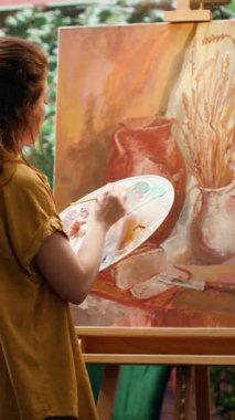Güzel sanatlar atölyesinde yarattığı sanat eseri için suluboya boya kullanan ve paletteki renkleri karıştıran dikey video kadın ressam. Tuvale kırsal öğeler çizen kadın, atölyede yaratıcı süreç