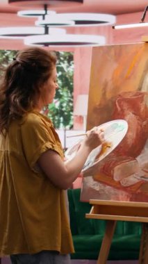 Güzel sanatlar atölyesinde yarattığı sanat eseri için suluboya boya kullanan ve paletteki renkleri karıştıran dikey video kadın ressam. Tuvale kırsal öğeler çizen kadın, atölyede yaratıcı süreç