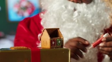 Noel Baba ev yapımı zencefilli kurabiye evini şekerleme ve boncuklarla süslüyor, Kuzey Kutbu kabin atölyesi. Siyah Noel Baba, tarçınlı tatlı pişirme geleneğini devam ettiriyor. Kamera A.