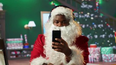 Neşeli Aziz Nicholas, Noel şarkılarını cep telefonundan dinliyor, takım elbiseli siyah Noel Baba kış tatili ruhunu kütük gibi kabinine yayıyor. Dijital teknoloji. Kamera A.