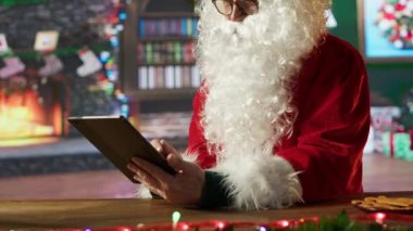 Noel Baba, tatil süslenmiş Kuzey Kutbu kütük kulübesinde internet sayfalarında gezinmek için bir tablet kullanıyor. Bayram ve Noel geleneklerinin tadını çıkarıyorum, hediye teslimatı hazırlıyorum. Kamera A.