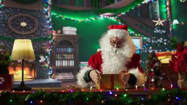 Noel Baba, Noel Baba 'nın Noel süslemeleriyle çevrili olan Kuzey Kutbu atölyesinde, festival kağıdı ve kurdelelerle hediye kutusunu paketliyor. Noel Baba kış kutlaması için sürprizler hazırlar. Kamera B.