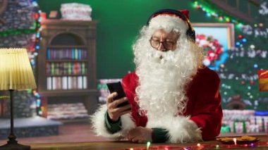 Neşeli Aziz Nicholas cep telefonundan Noel şarkıları dinliyordu. Beyaz sakallı adam kış tatili ruhunu odasındaki kulübesine yayıyordu. Dijital teknoloji. Kamera A.