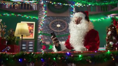 Neşeli Aziz Nicholas cep telefonundan Noel şarkıları dinliyordu. Beyaz sakallı adam kış tatili ruhunu odasındaki kulübesine yayıyordu. Dijital teknoloji. Kamera B.