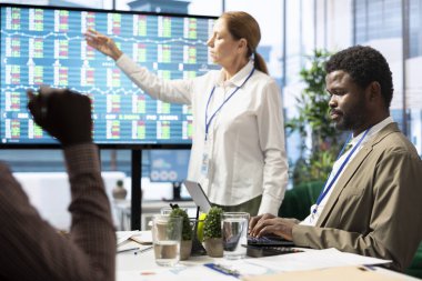 Genel müdür interaktif panoda bilgi grafikleri ve satış numaralarıyla bir sunum yapıyor, personele organizasyonel ilerlemeyi gösteriyor. Çalışanlar analiz planlarını gözden geçiriyorlar.